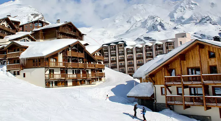Station de ski enneigée avec chalets en bois, montagnes en arrière-plan et skieurs sur les pistes.