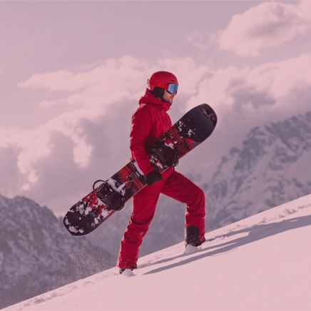 Un snowboarder en combinaison rouge marche dans la neige, portant sa planche sur une pente enneigée, avec des montagnes en arrière-plan.