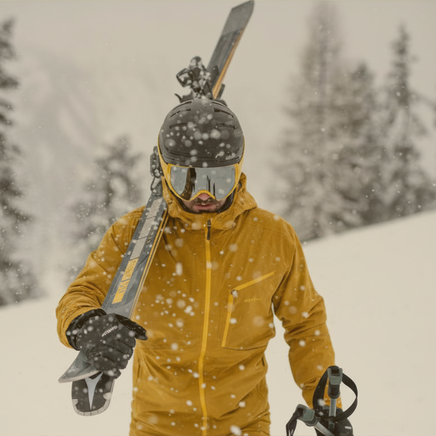 Un skieur en veste jaune et casque, portant ses skis sur l'épaule, sous une forte chute de neige en montagne.
