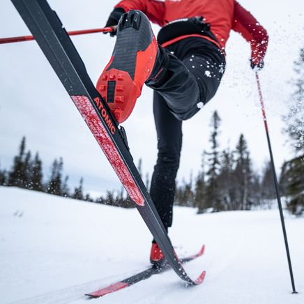 Vue en contre-plongée d'un skieur de fond en action, sa botte rouge sur un ski Atomic noir, projetant de la neige sur la piste.