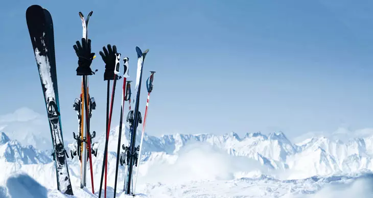 Skis, bâtons et gants dressés dans la neige devant un paysage de montagnes enneigées sous un ciel bleu.