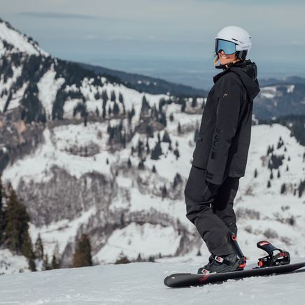 Un snowboarder en tenue noire avec casque blanc et lunettes bleues, debout sur sa planche sur une piste enneigée avec montagnes en arrière-plan.