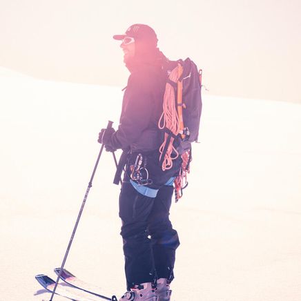 Un skieur de randonnée souriant, avec sac à dos, corde et piolets, dans un paysage de montagne enneigé et lumineux.