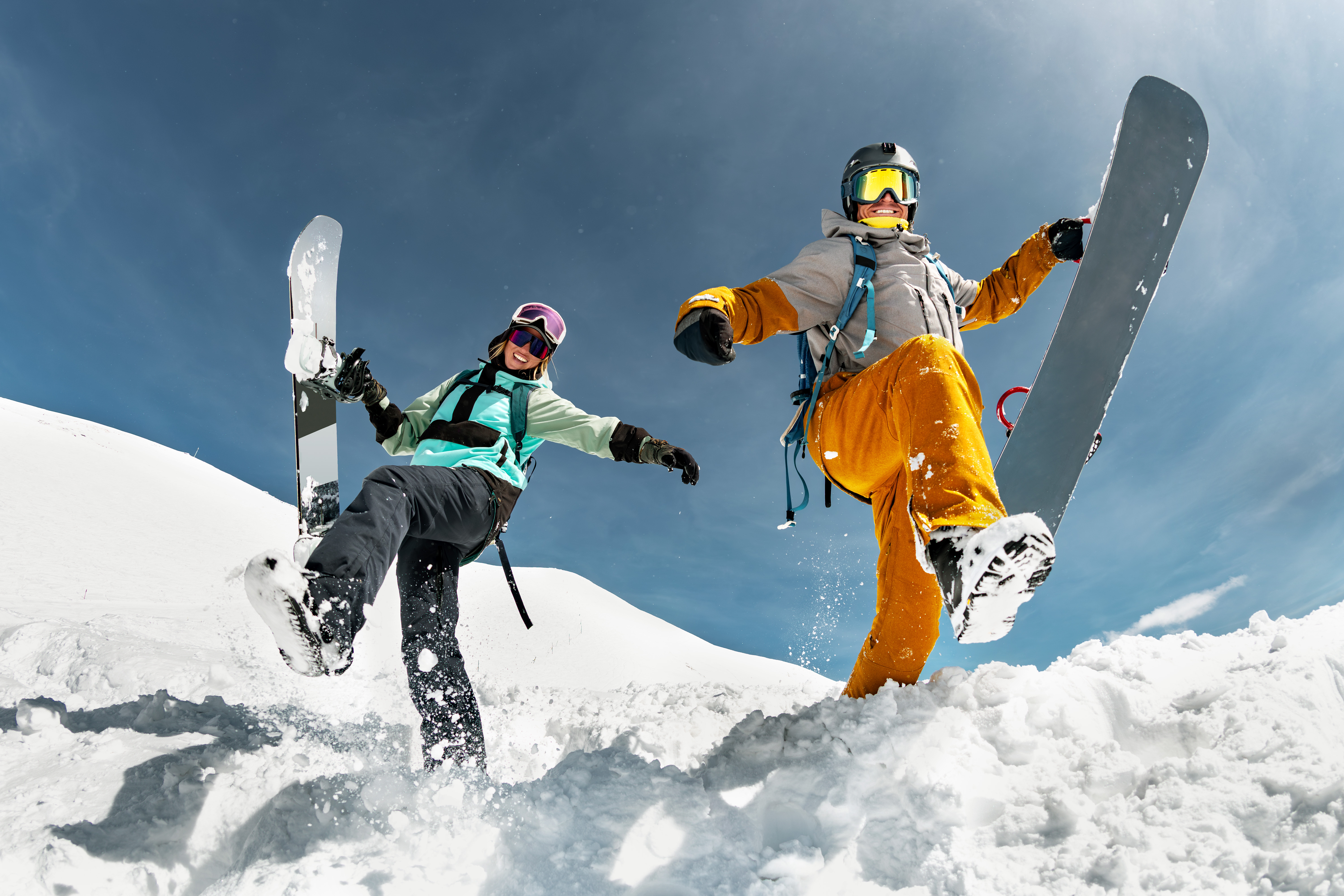 Deux snowboarders joyeux dans la neige, levant une jambe en l'air, sous un ciel bleu.