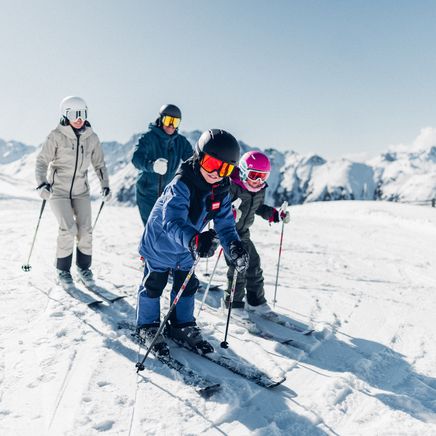 Une famille de quatre personnes skie sur une piste ensoleillée avec des montagnes enneigées en arrière-plan.