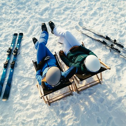 Deux skieurs se détendent dans des chaises longues sur une piste enneigée, avec leurs skis posés à côté. Vue plongeante.