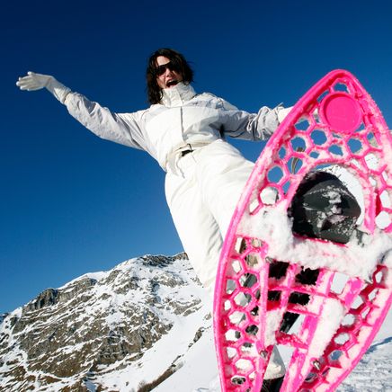 Femme enthousiaste en blanc, lunettes, bras écartés, avec une raquette à neige rose vif sur une pente enneigée sous un ciel bleu.
