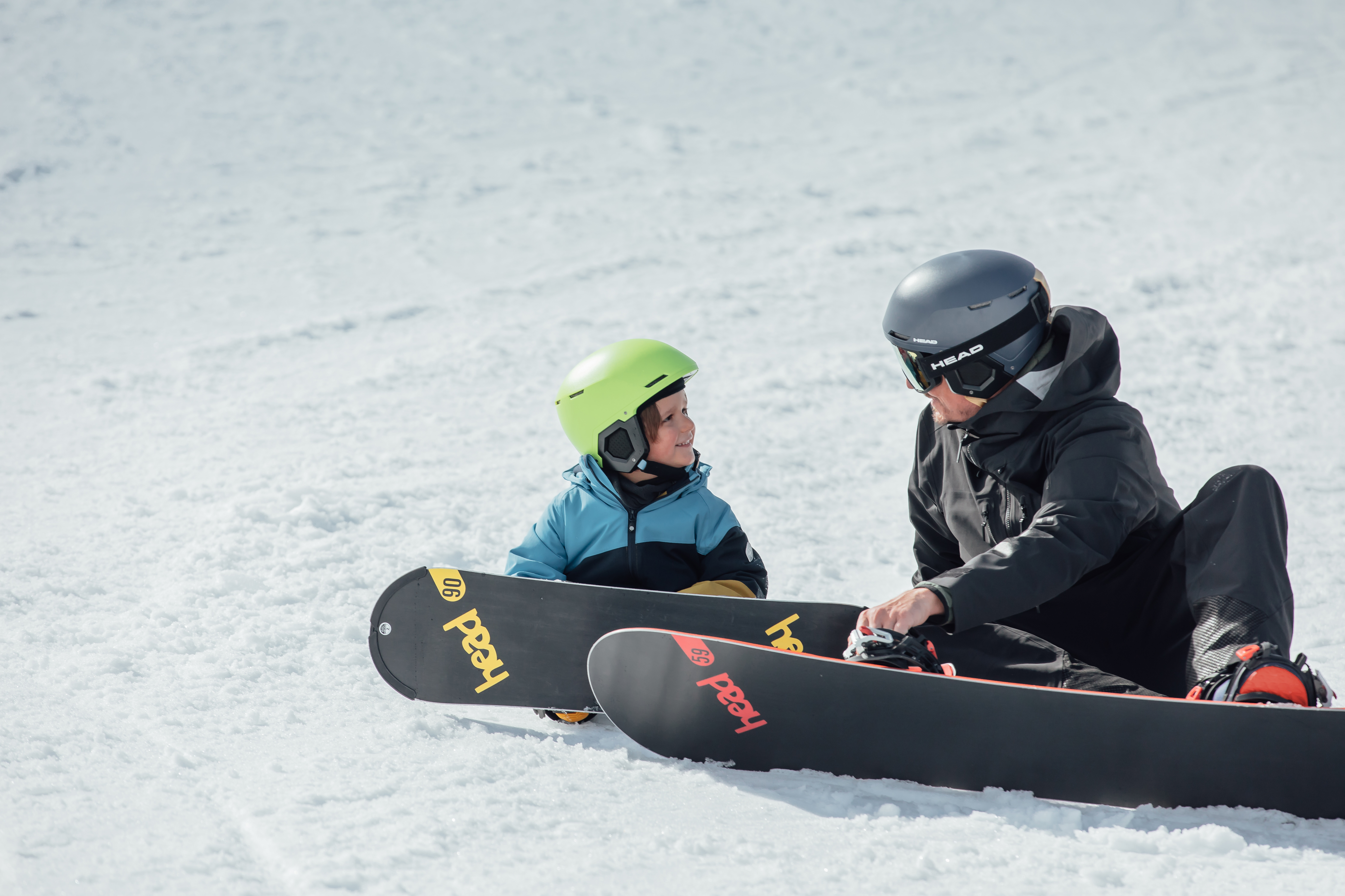 Un adulte ajuste sa fixation de snowboard tandis qu'un enfant casqué et souriant le regarde attentivement sur une piste enneigée.