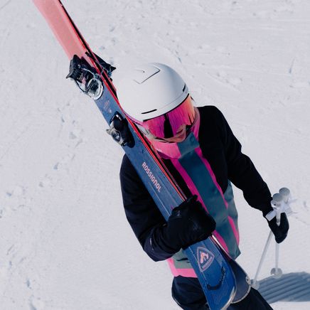 Skieur portant casque blanc et lunettes roses, skis Rossignol bleus sur l'épaule et bâtons, marchant dans la neige.