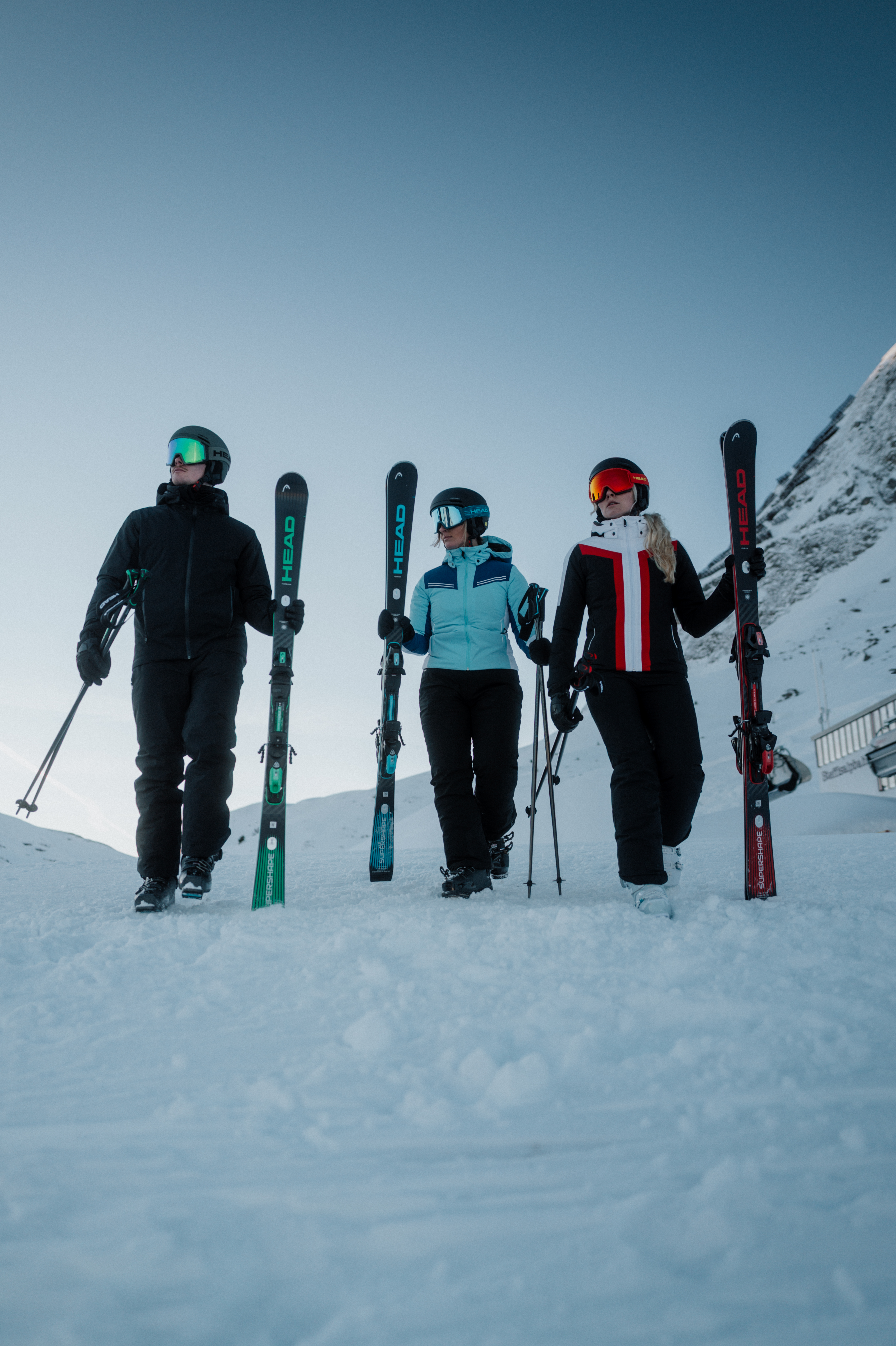 Trois skieurs, un homme et deux femmes, équipés de skis et bâtons, posent sur une montagne enneigée sous un ciel clair.