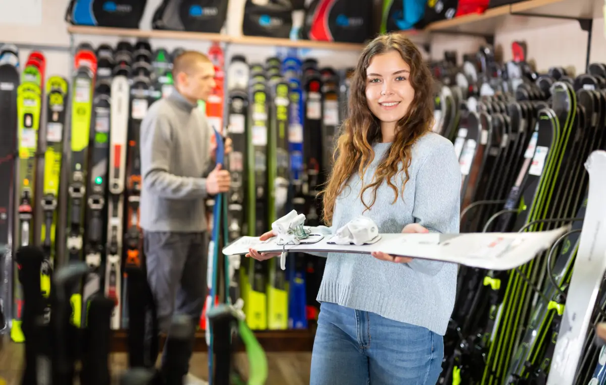 Jeune femme souriante tenant une planche de ski avec fixations dans un magasin de sport, des skis sont exposés en arrière-plan.