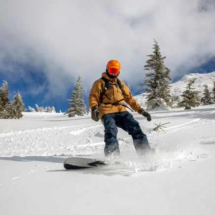 Un snowboarder avec un casque orange dévale une pente enneigée, soulevant de la poudreuse sous un ciel bleu et des sapins.