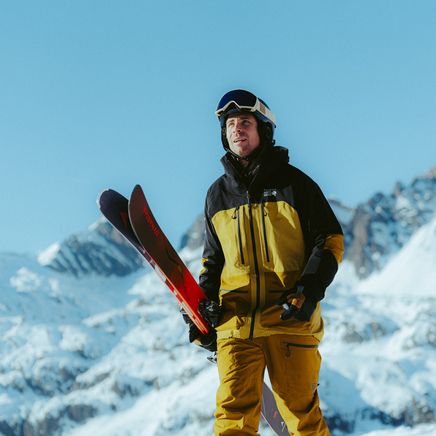 Un skieur en tenue jaune et noire, casque et lunettes, portant ses skis dans un paysage montagnard enneigé sous un ciel bleu clair.