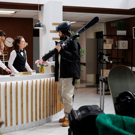 Des clients de sports d'hiver arrivent à la réception d'un hôtel, l'un portant des skis, l'autre regardant son téléphone près d'un snowboard.