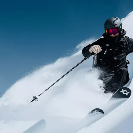 Un skieur en tenue noire dévale une pente enneigée, créant un nuage de poudreuse sous un ciel bleu éclatant.