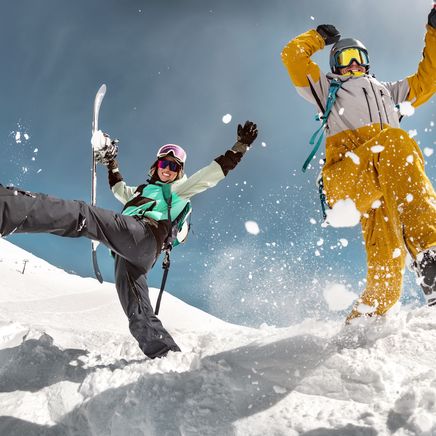 Skieur et snowboarder, bras levés et joyeux, s'amusent dans la neige, des flocons virevoltant sous un ciel bleu éclatant.