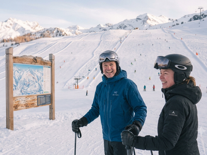 Deux skieurs souriants devant un plan des pistes, avec un domaine skiable et des montagnes enneigées en arrière-plan.