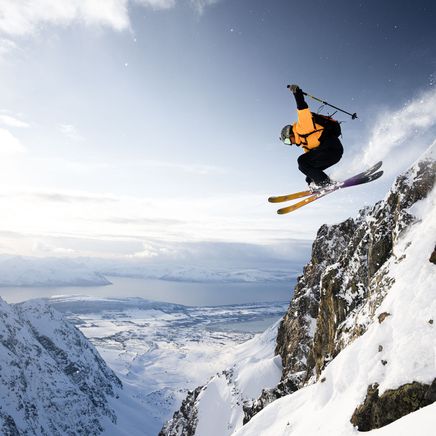 Skieur freeride en veste orange en plein vol après un saut d'une corniche rocheuse enneigée, vue sur un fjord et des montagnes.
