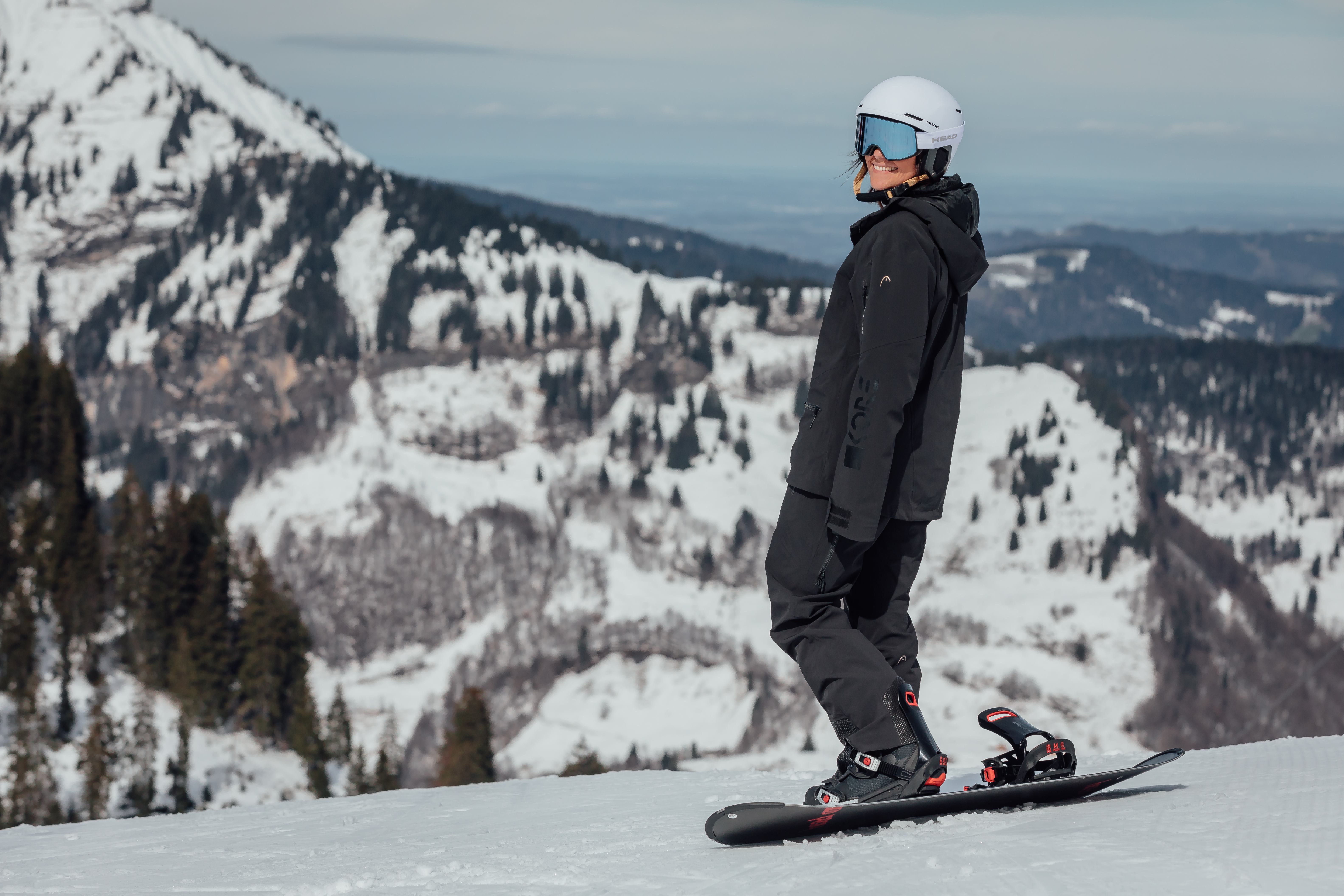 Une personne en tenue de snowboard noire, casque blanc et lunettes bleues, se tient sur une planche à neige sur une piste de montagne enneigée.