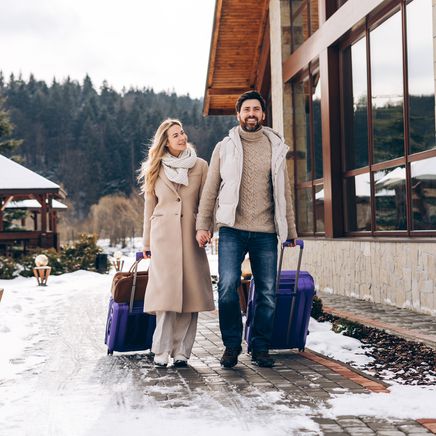 Un couple souriant arrive avec ses valises devant un chalet de luxe enneigé en hiver.