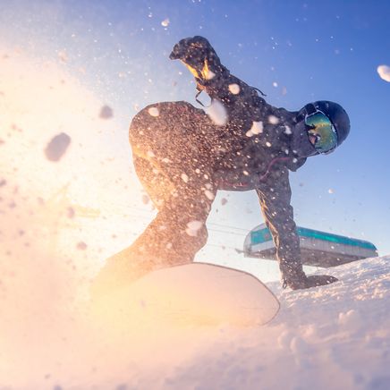 Snowboardeur en plein virage sur neige poudreuse, éclaboussures illuminées par le soleil rasant. Télécabine en arrière-plan.