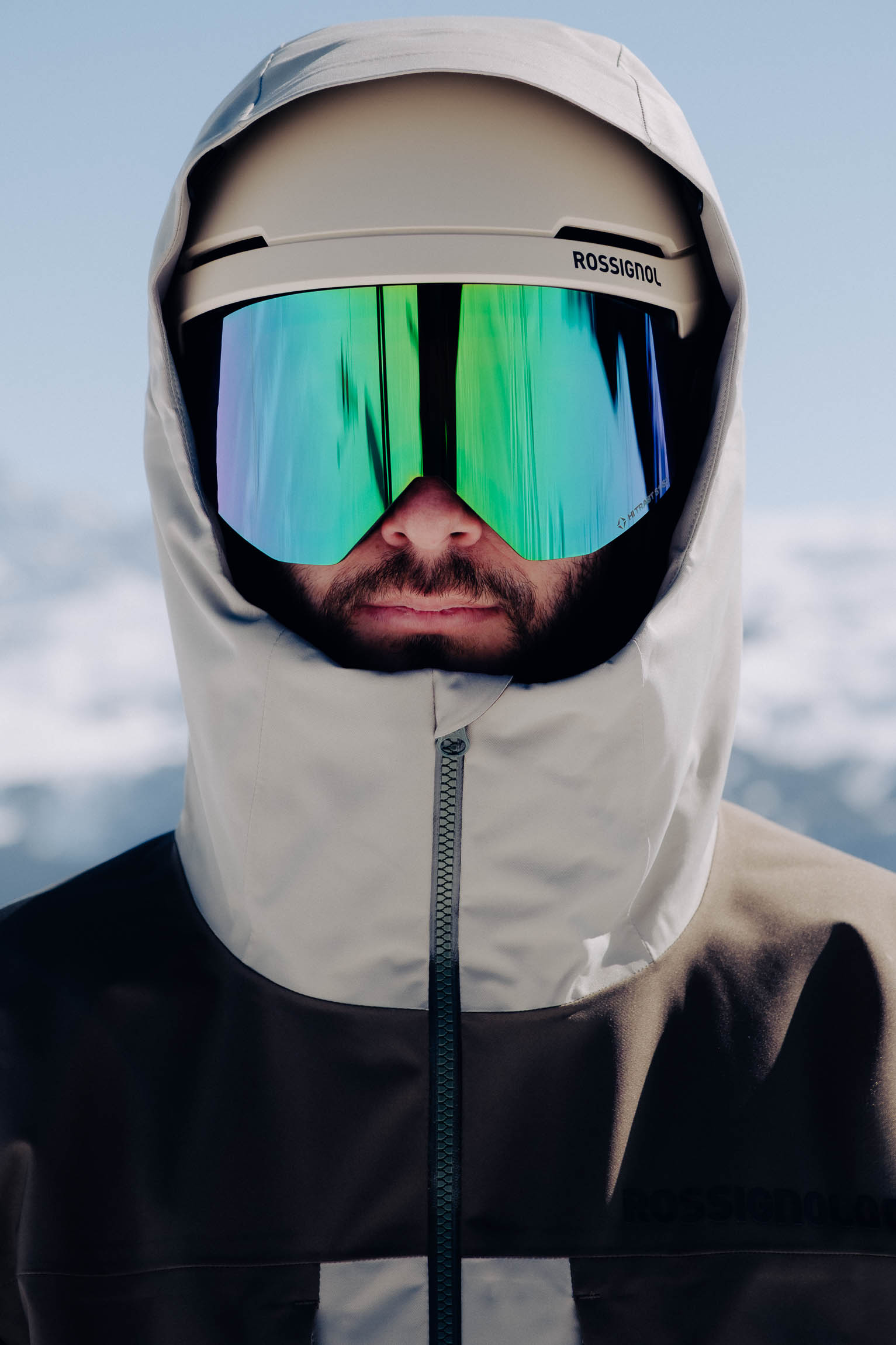 Vue rapprochée d'un homme barbu en équipement de ski : casque Rossignol crème, masque de ski réfléchissant vert/bleu, veste à capuche.
