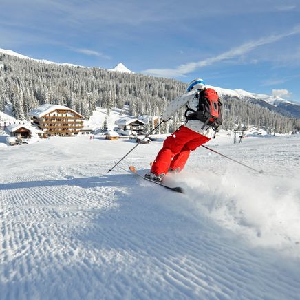 Un skieur en rouge dévale une piste enneigée sous un ciel bleu, avec des chalets et une forêt de pins en arrière-plan.