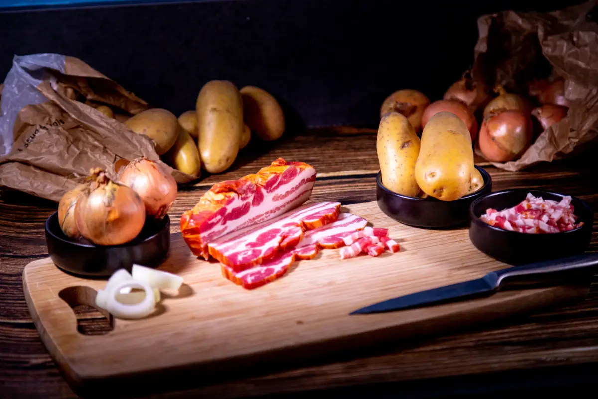 Ingrédients pour cuisiner : lard fumé en tranches et lardons, oignons, et pommes de terre sur une planche à découper avec un couteau.